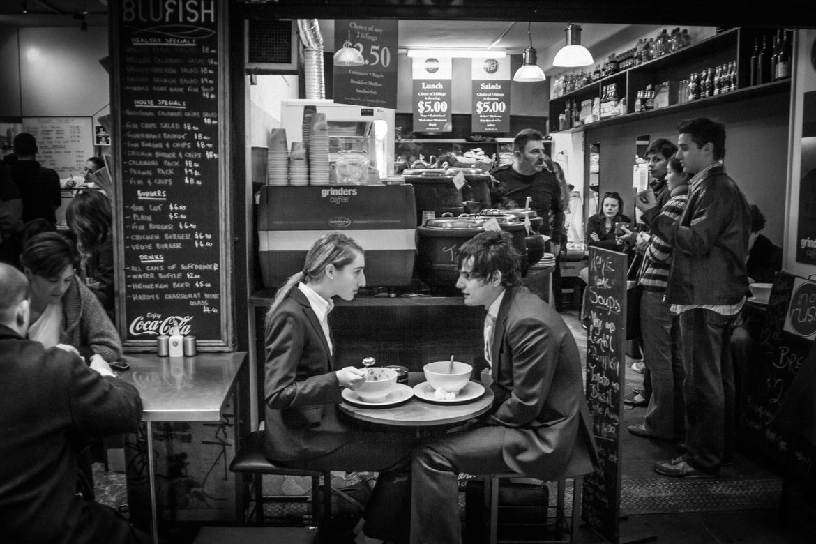 A black-and-white interior of a crowded café. Two people in dark clothing sit facing each other at a small round table with bowls, while other customers and staff fill the background. Chalkboard menus and warm overhead lights define the space.
