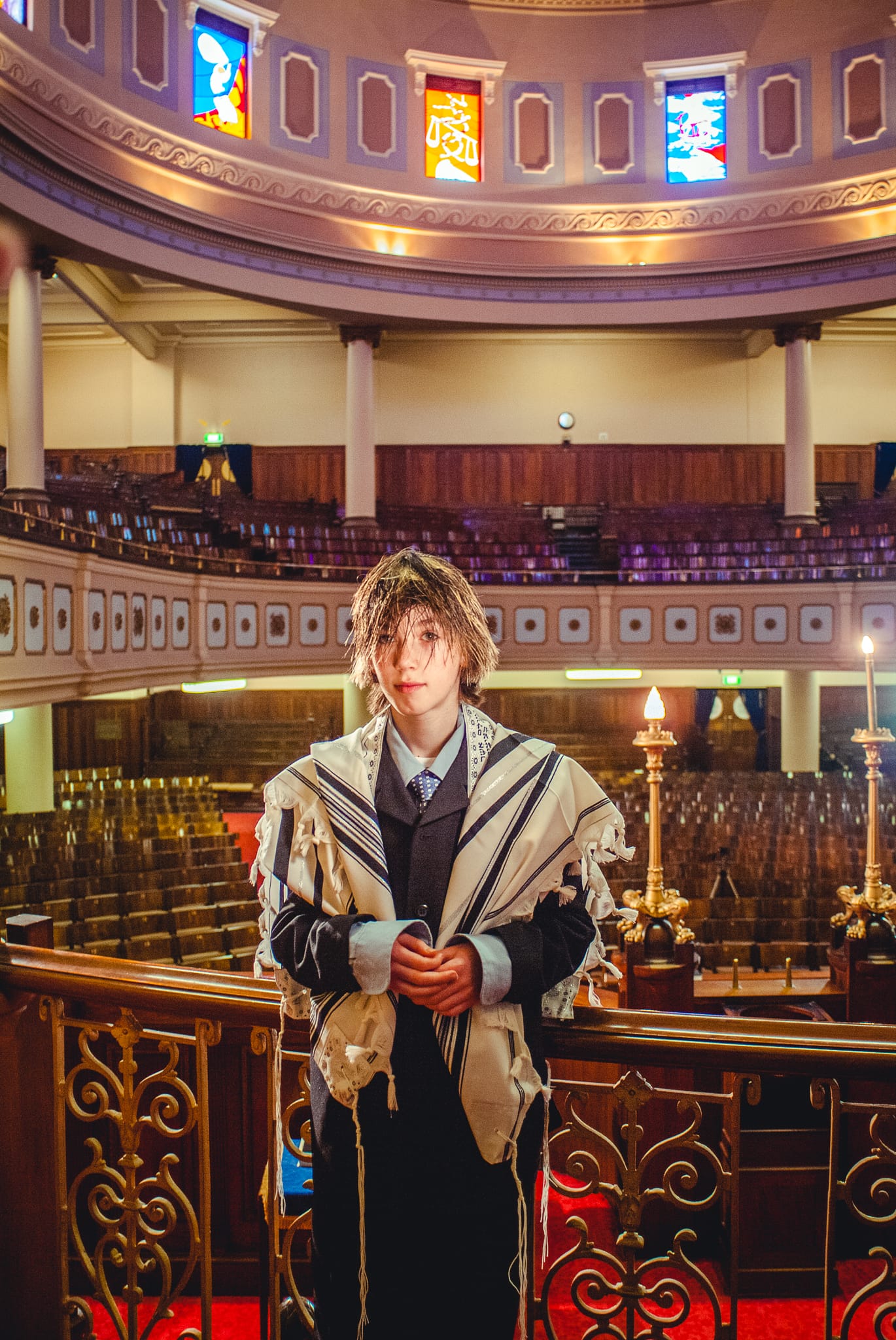 Colour milestone portrait of a Bar Mitzvah boy standing alone in a grand synagogue beneath stained glass windows and golden candelabra — Arnold Szmerling Visual Legacy Artist