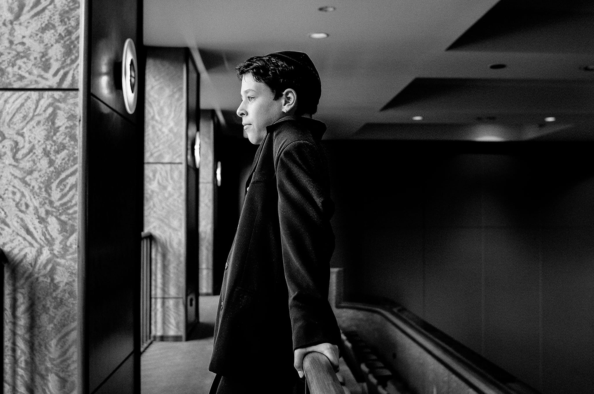 Cinematic black and white profile portrait of a Bar Mitzvah boy in a dark coat at a grand venue escalator — Arnold Szmerling Visual Legacy Artist