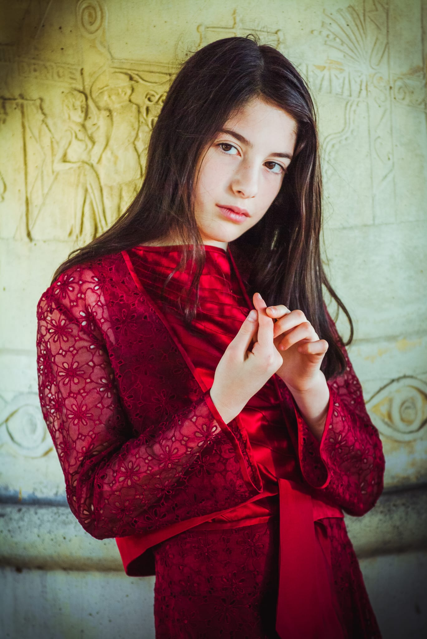 Colour milestone portrait of a young girl in a crimson lace dress standing against ancient carved stone relief — Arnold Szmerling Visual Legacy Artist