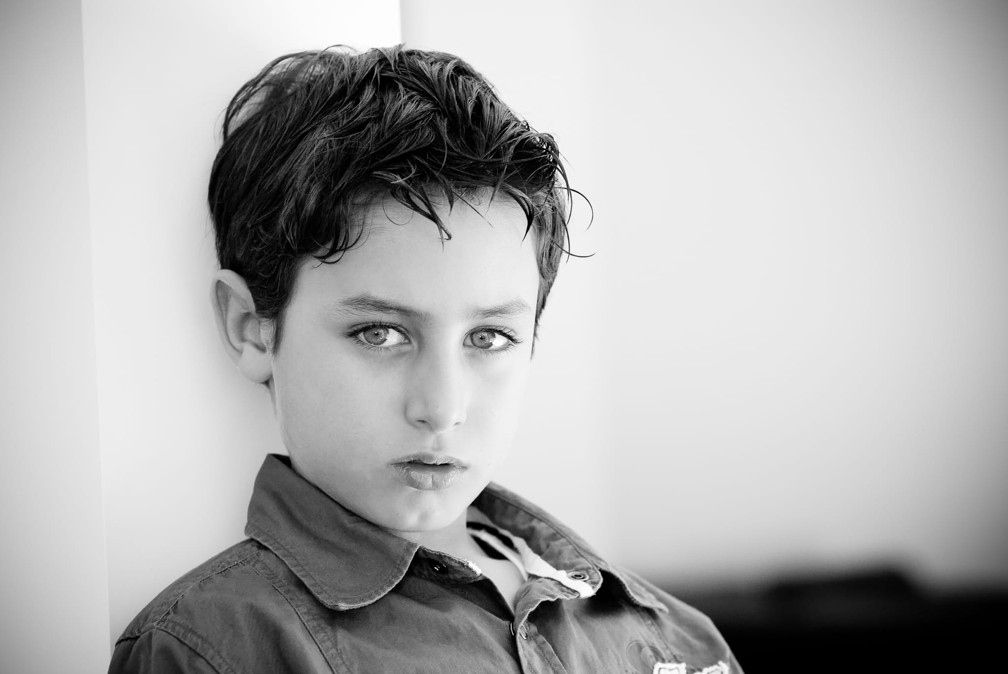 Cinematic black and white close portrait of a young boy with expressive eyes leaning against a white wall — Arnold Szmerling Visual Legacy Artist
