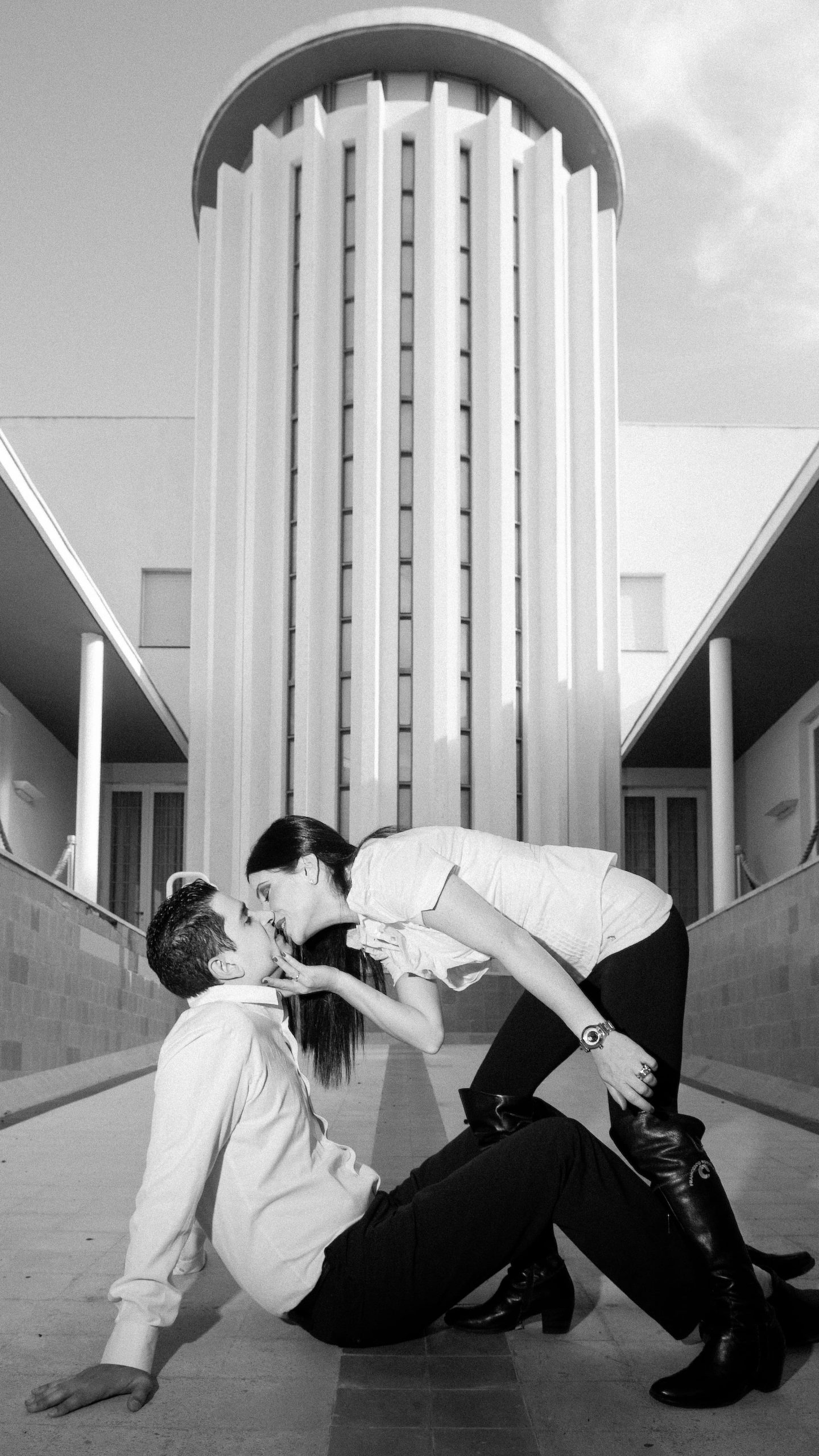 Cinematic black and white portrait of a couple sharing a kiss against a monumental modernist building — Arnold Szmerling Visual Legacy Artist