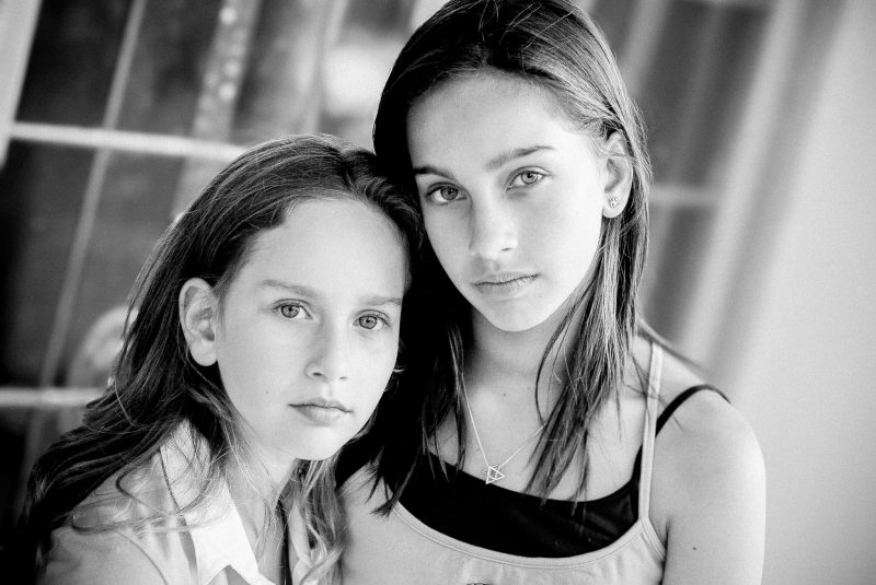 Intimate black and white portrait of two sisters pressed together gazing directly at camera in natural window light — Arnold Szmerling Visual Legacy Artist