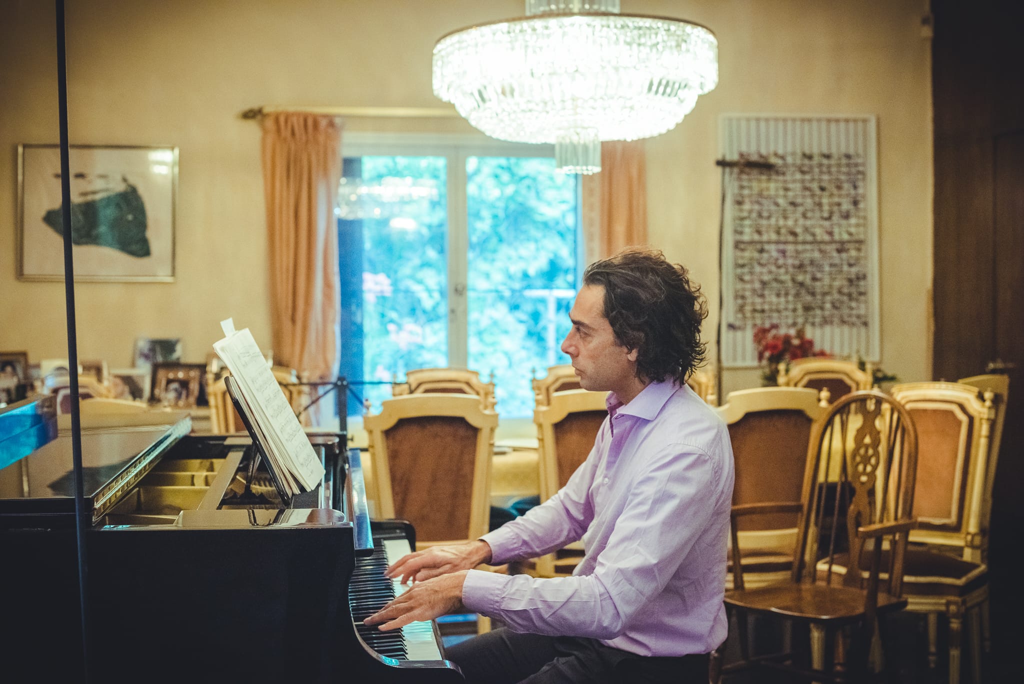A man absorbed in playing a grand piano beneath a crystal chandelier in a formal dining room — Arnold Szmerling Visual Legacy Artist