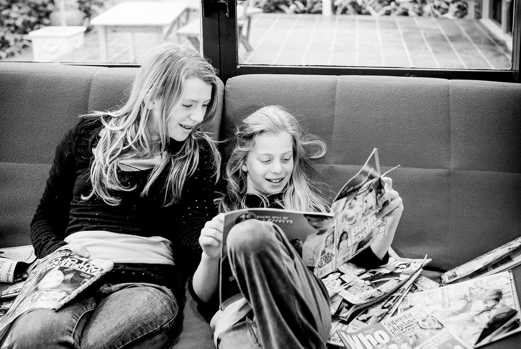 Two sisters sharing a quiet, joyful moment with magazines at home in Melbourne – candid family connection by Arnold Szmerling