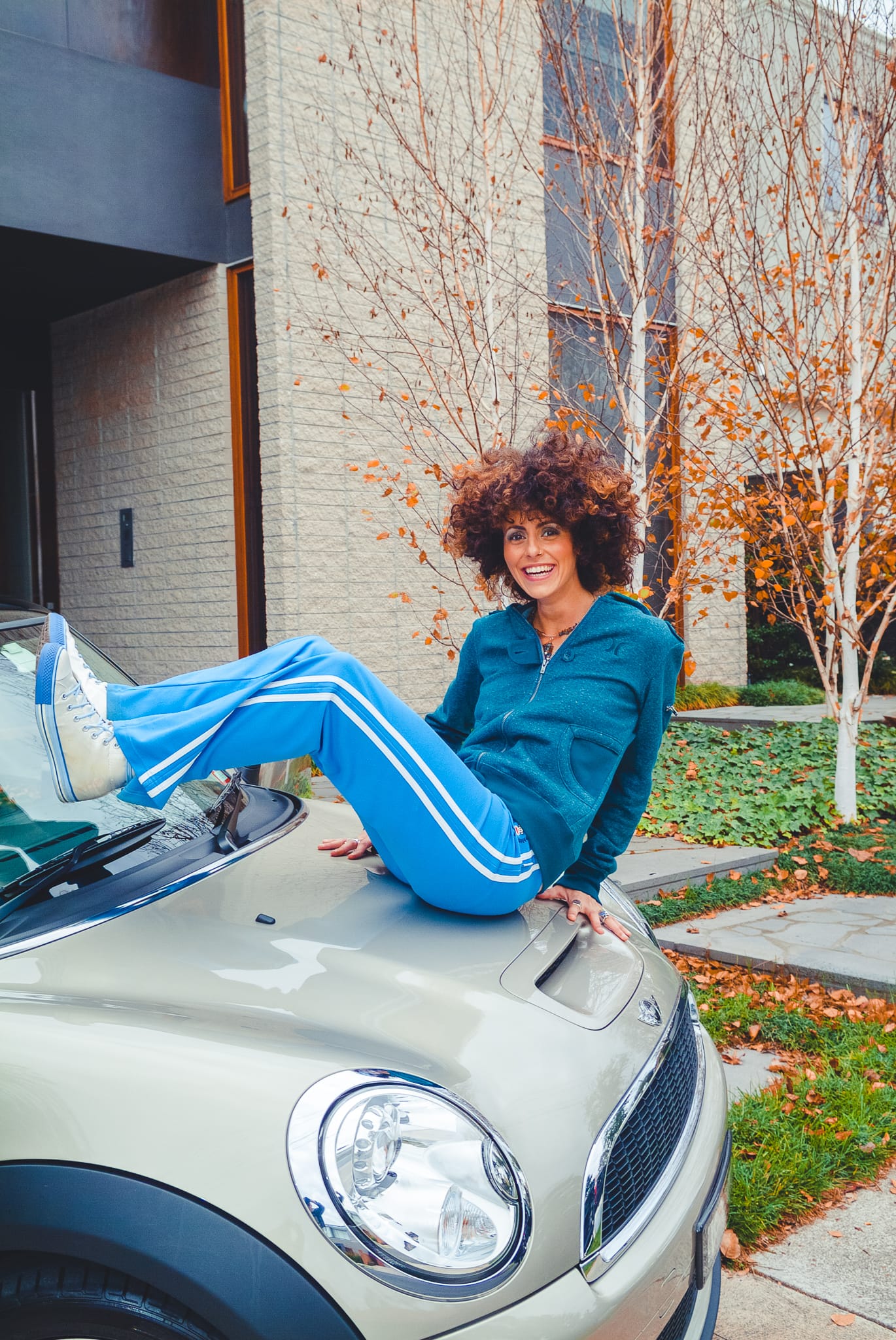 A woman in a blue tracksuit laughing on the hood of a Mini Cooper in an autumn street setting — Arnold Szmerling Visual Legacy Artist